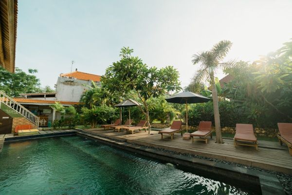Serene tropical infinity pool at a Balinese resort, with wooden lounge chairs, black umbrellas, palm trees, and lush gardens under a sunny sky. - Bali Villas